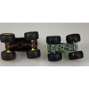 May include: Two toy monster trucks, one brown and one green, are displayed upside down. The brown truck has yellow wheels, while the green truck has silver and green details. Both have large black tires.