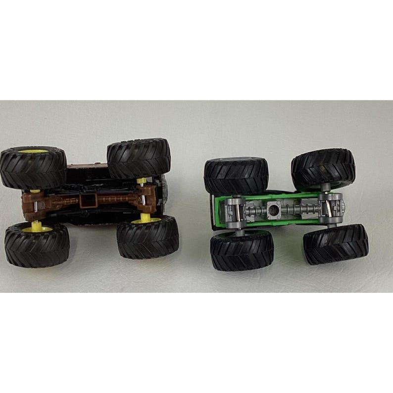 May include: Two toy monster trucks, one brown and one green, are displayed upside down. The brown truck has yellow wheels, while the green truck has silver and green details. Both have large black tires.