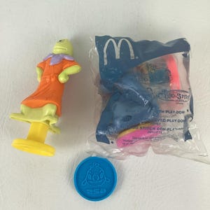 May include: A collection of McDonald's Happy Meal toys featuring characters from the Disney movie Lilo & Stitch. Includes a green and orange figure, a blue plastic coin, and a sealed bag with a Stitch toy and Play-Doh.