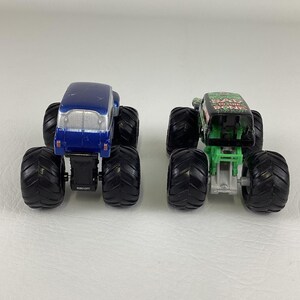 May include: Two toy monster trucks. One is blue with a silver top, the other is green and black with the text "BAD TO THE BONE". Both have large black tires and are designed for play.