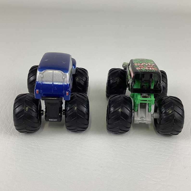 May include: Two toy monster trucks. One is blue with a silver top, the other is green and black with the text "BAD TO THE BONE". Both have large black tires and are designed for play.