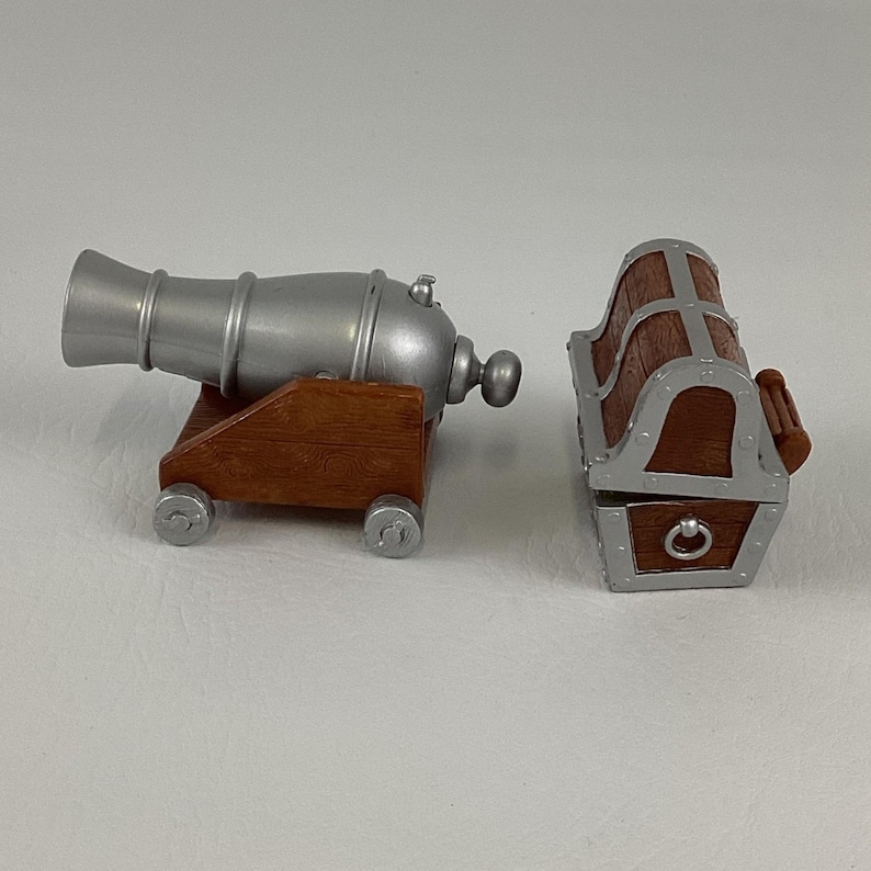 May include: A miniature toy cannon with a silver barrel and a brown wooden base. Beside it is a small treasure chest with a brown wooden body and silver accents. The chest has a hinged lid and a circular clasp.