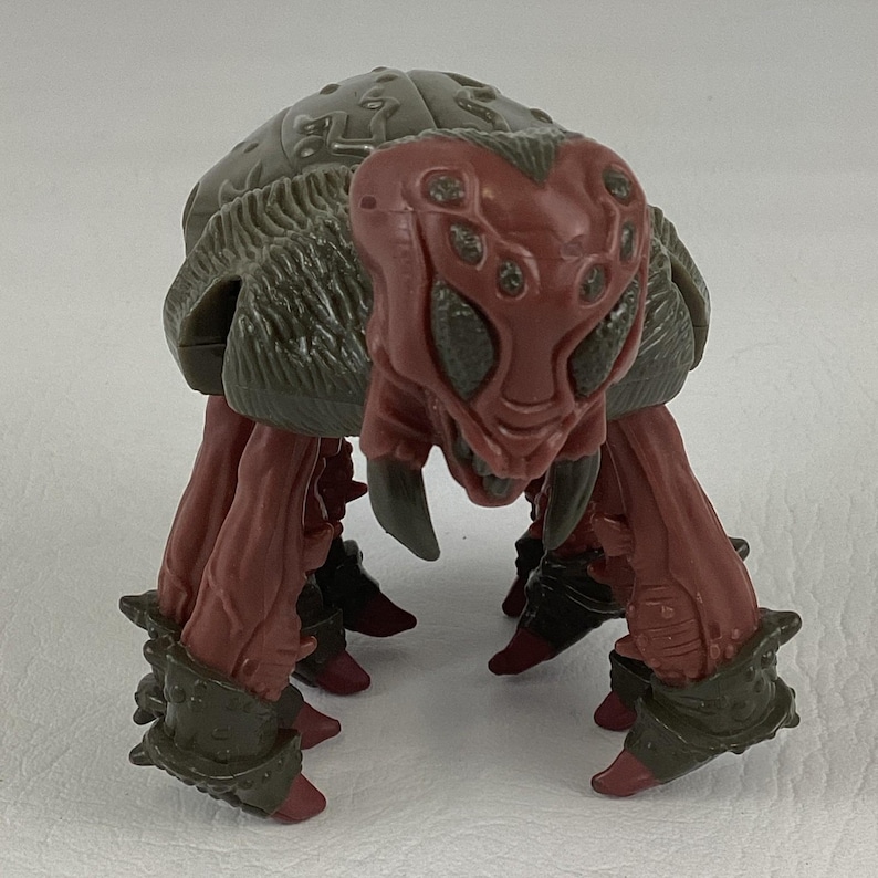 K&ouml;nnte beinhalten: Eine Kunststoff-Actionfigur eines mehrbeinigen Wesens mit dunkelrotem Kopf und Torso, mehreren Augen und scharfen Mandibeln. Das Wesen hat einen strukturierten olivgr&uuml;nen Panzer auf dem R&uuml;cken und passende olivgr&uuml;ne R&uuml;stungsteile an den Beinen.