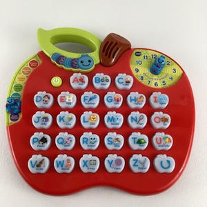 May include: A red and green VTech ABC Learning Apple toy. The toy is shaped like an apple and has buttons for each letter of the alphabet, a clock, and a caterpillar. Each button has a picture and word.