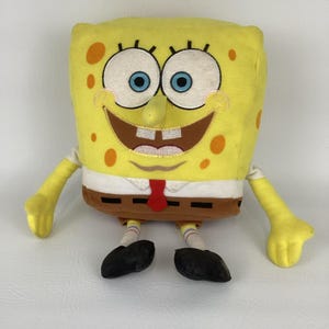 May include: A Spongebob Squarepants plush toy. The yellow sponge character has large blue eyes, a wide smile, and orange spots. He wears a white shirt, brown trousers, a red tie, and black shoes. The toy is designed to resemble the character from the cartoon.