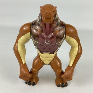 May include: A brown and cream-colored plastic action figure of a reptilian creature. The figure features a clear plastic chest plate revealing a dark red internal structure, muscular limbs, a wide stance, and black clawed feet and hands.