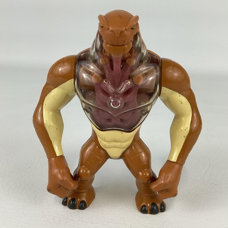 May include: A brown and cream-colored plastic action figure of a reptilian creature. The figure features a clear plastic chest plate revealing a dark red internal structure, muscular limbs, a wide stance, and black clawed feet and hands.