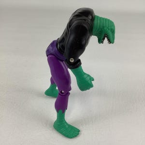 Puede incluir: Una figura de acci&oacute;n de pl&aacute;stico con cabeza de reptil verde y boca abierta, torso negro, pantalones morados y manos y pies verdes. La figura est&aacute; en una postura encorvada e inclinada hacia adelante con articulaciones en hombros y caderas.