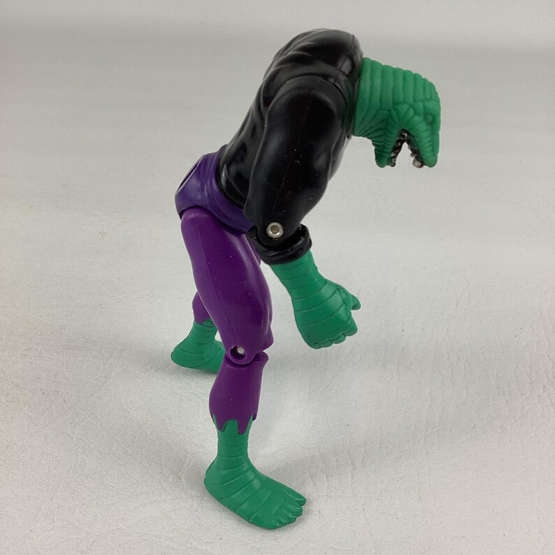 Puede incluir: Una figura de acci&oacute;n de pl&aacute;stico con cabeza de reptil verde y boca abierta, torso negro, pantalones morados y manos y pies verdes. La figura est&aacute; en una postura encorvada e inclinada hacia adelante con articulaciones en hombros y caderas.