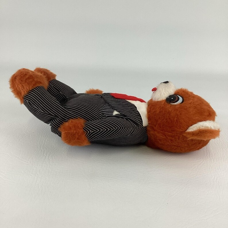 Pode incluir: Uma raposa de pel&uacute;cia vintage vestindo um terno de riscas preto e branco com uma gravata vermelha. A raposa tem pele laranja, detalhes brancos e olhos grandes de desenho animado. O brinquedo est&aacute; deitado de costas.