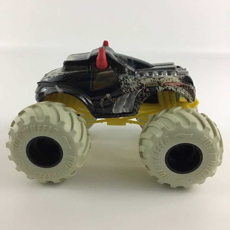 May include: Black monster truck toy with red horns, yellow wheels, and a white tire tread. The truck has a camouflage pattern and the words "Monster Trucks" on the side.