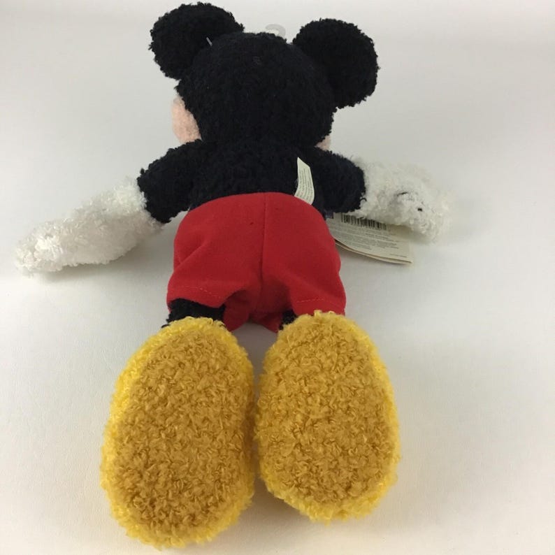 May include: A plush Mickey Mouse toy, viewed from the back, with black fur, red shorts, and yellow feet. The toy has white gloves and a pink face. A white tag is attached to the toy.