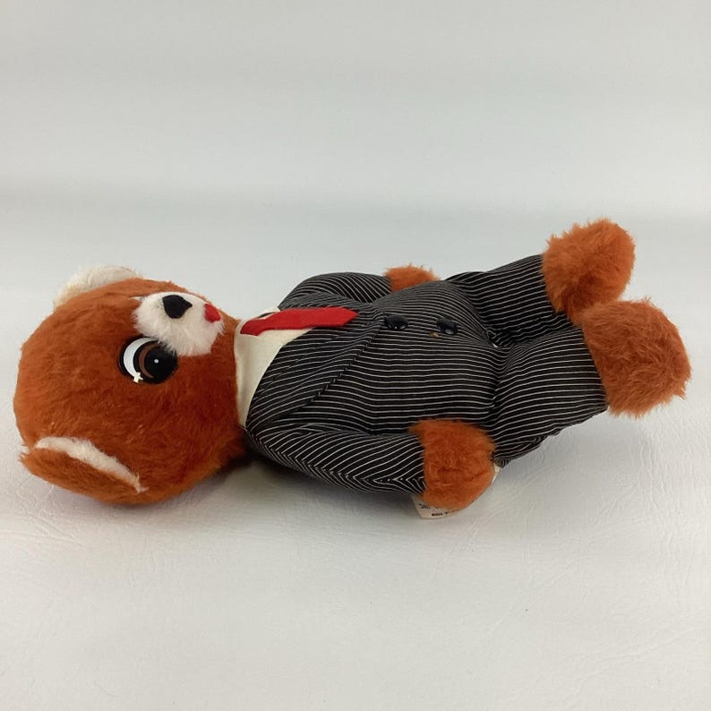 Pode incluir: Um brinquedo de urso de pel&uacute;cia vintage vestindo um terno de riscas de giz preto com gravata vermelha e camisa branca. O urso tem pelo marrom com detalhes brancos e patas laranja. O brinquedo est&aacute; deitado de costas.