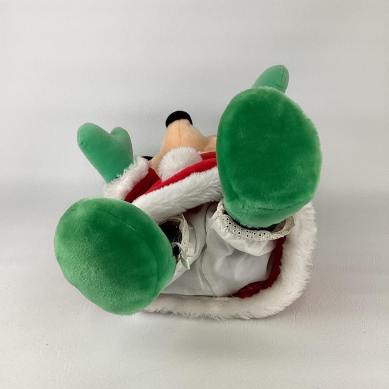 Puede incluir: Un juguete de peluche con un traje estilo Santa Claus rojo y blanco con ribete de piel sint&eacute;tica blanca. El personaje tiene pies y manos de peluche de color verde brillante, una cara de color canela y pantalones de tela blanca con encaje.