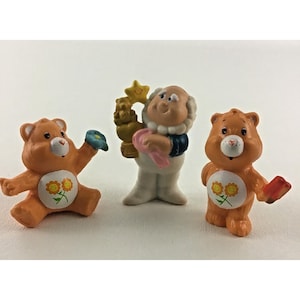 May include: Two orange bear figurines with white belly patches featuring two yellow flowers and green stems, one sitting and one standing. Between them stands a light-skinned character with a white outfit, holding a gold star trophy.