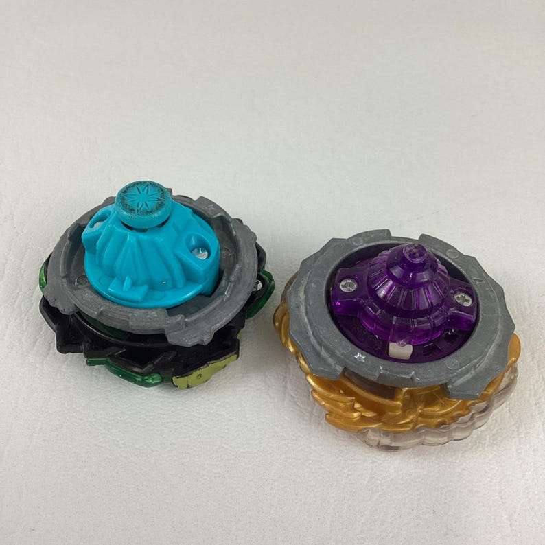 May include: Two colorful spinning tops. One has a teal top with a gray and black base. The other has a purple top with a gray and gold base. The tops are designed with multiple layers and intricate details.