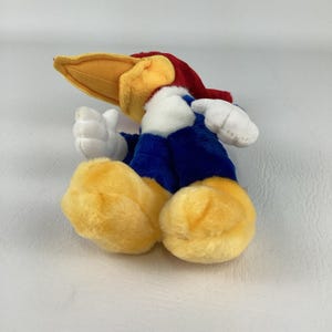 Op de afbeelding: Een pluche speelgoed van Woody Woodpecker, met een gele snavel, rode hoofdkleur en blauwe lichaam. Het speelgoed heeft witte handschoenen en gele voeten en ligt op zijn zij.