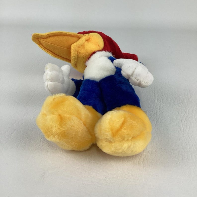 Op de afbeelding: Een pluche speelgoed van Woody Woodpecker, met een gele snavel, rode hoofdkleur en blauwe lichaam. Het speelgoed heeft witte handschoenen en gele voeten en ligt op zijn zij.