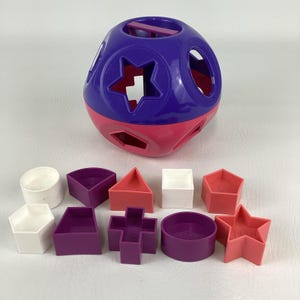 Puede incluir: Un juguete clasificador de formas colorido con una bola morada y rosa con varias formas recortadas. Incluye diez formas geométricas diferentes en blanco, morado y rosa. Un juguete educativo clásico para niños pequeños.