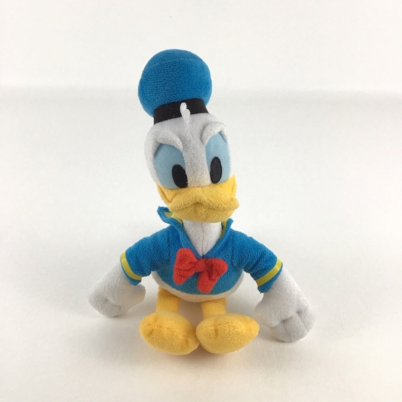 May include: A plush toy of Donald Duck sitting down. He is wearing a blue sailor shirt with a red bow tie and a blue sailor hat. He has a yellow beak and white feathers.