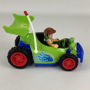 May include: A green toy car with a Woody figure inside. The car has black tires with red rims, a blue bumper, and a white pole with a red tip. A blue water splash design is on the side.