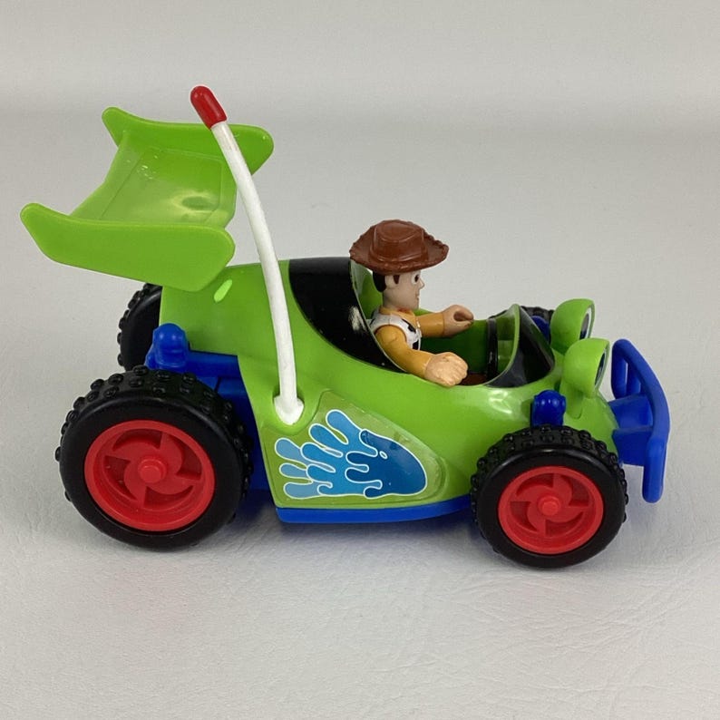 May include: A green toy car with a Woody figure inside. The car has black tires with red rims, a blue bumper, and a white pole with a red tip. A blue water splash design is on the side.