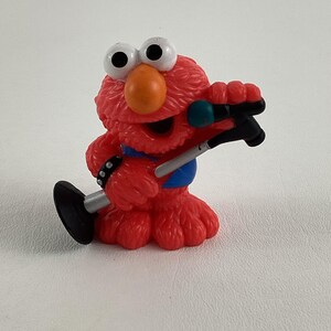 Puede incluir: Figura de juguete Elmo roja sosteniendo un soporte de micrófono. Elmo lleva una camisa azul y blanca con una banda negra tachonada alrededor de la muñeca.