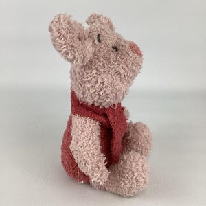 May include: A plush toy pig in a seated position. The pig is primarily light pink with a darker pink vest and nose. The toy has a textured, fuzzy appearance and small black eyes.