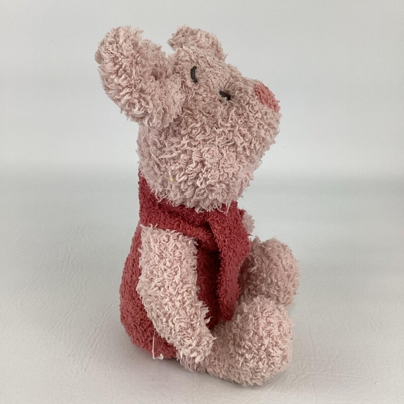 May include: A plush toy pig in a seated position. The pig is primarily light pink with a darker pink vest and nose. The toy has a textured, fuzzy appearance and small black eyes.