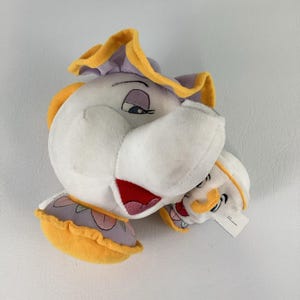 Può includere: Un set di peluche composto da una grande teiera bianca con coperchio e base a balze viola e gialle, insieme a una tazzina bianca più piccola con bordo e base gialli, entrambi con tratti del viso ricamati e dettagli rossi sulla bocca.