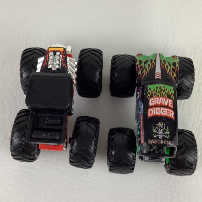 May include: Two toy monster trucks, one red and black, the other black and green. The red truck has silver accents and the black truck has "Grave Digger" graphics and a skull design. Both have large black tires.