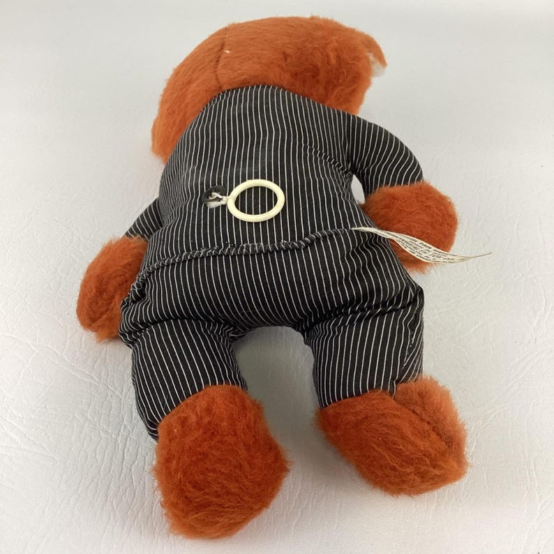 Pode incluir: Um urso de pel&uacute;cia vintage, visto por tr&aacute;s, com p&ecirc;lo laranja e um fato de riscas pretas e brancas. O urso tem patas laranja e uma cabe&ccedil;a laranja a condizer. Um anel de pl&aacute;stico branco est&aacute; preso nas costas do fato.