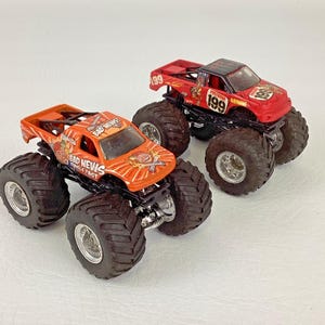 Puede incluir: Dos monster trucks de juguete. Uno es naranja con el texto "BAD NEWS TRAVELS FAST" y un gr&aacute;fico de dibujos animados. El otro es rojo con el n&uacute;mero "199" y el texto "PRESTADOR RACING". Ambos tienen neum&aacute;ticos negros grandes.