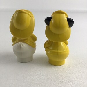 May include: Two yellow plastic figurines of ducks wearing hats. One duck has a white body and the other duck has a yellow body.