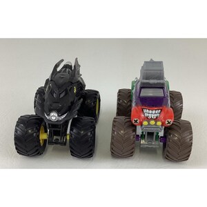 May include: Two toy monster trucks. One is black with yellow accents, resembling a bat. The other is red, purple, and green, with a grinning face design. Both have large, black tires.