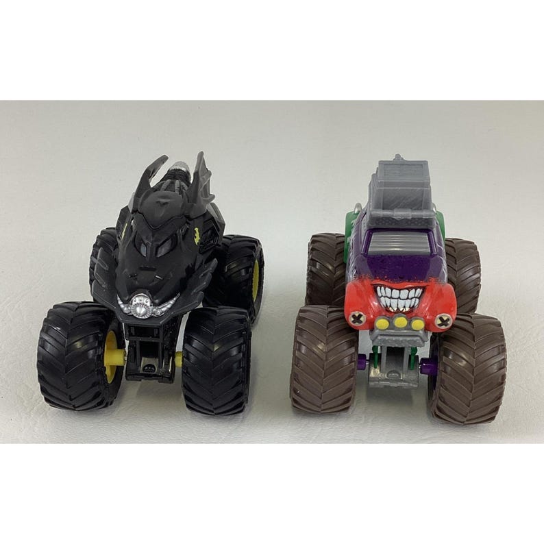 May include: Two toy monster trucks. One is black with yellow accents, resembling a bat. The other is red, purple, and green, with a grinning face design. Both have large, black tires.