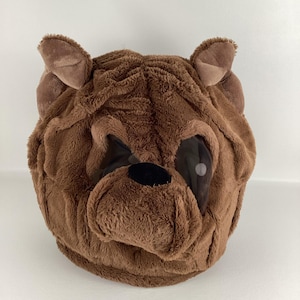 Puede incluir: Un disfraz de cabeza de oso de peluche marrón con aberturas para los ojos y una nariz negra. El disfraz tiene dos orejas marrones y está hecho de un material suave y difuso. Este disfraz es adecuado para Halloween o fiestas temáticas.