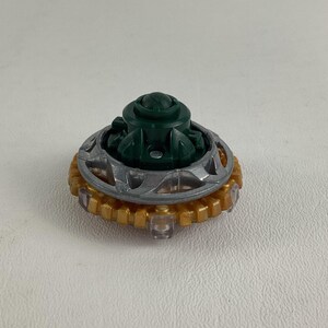 Puede incluir: Primer plano de una peonza de juguete. La peonza presenta un componente central verde oscuro, un anillo plateado y un anillo exterior dorado. El juguete est&aacute; dise&ntilde;ado para girar y jugar.