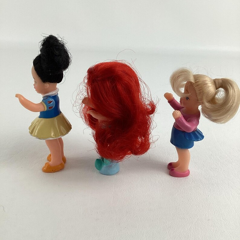 May include: Three small plastic figurines of cartoon characters. The first figure has black hair, a blue and yellow dress, and orange shoes. The second figure has red hair and a blue base. The third figure has blonde hair, a pink top, and a blue skirt.