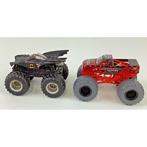 May include: Two toy monster trucks. One is black with a Batman logo, the other is red with the text "NORTHERN NIGHTMARE". Both have large, gray tires and are designed for off-road play.