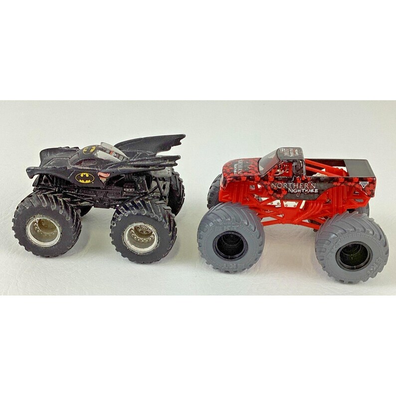 May include: Two toy monster trucks. One is black with a Batman logo, the other is red with the text "NORTHERN NIGHTMARE". Both have large, gray tires and are designed for off-road play.