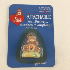 以下が含まれることがあります： ケアベアロゴと「ATTACHABLE Fun...festive... attaches to anything! (See other side)」というテキストが書かれた青と白のカード。カードには、お腹に黄色のカップケーキが付いた小さな茶色のシェアベアチャームが付いています。チャームは金色のチェーンに付いています。カードの下部には「SHARE BEARTM 53149」というテキストが印刷されています。