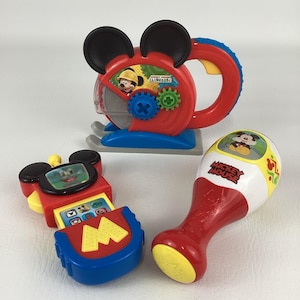 May include: Three colorful Mickey Mouse Clubhouse toys. The toys include a red and blue saw with Mickey Mouse, a red and blue phone with a screen, and a red and white maraca-like toy with Mickey Mouse.