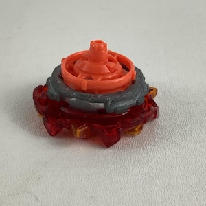 May include: A red and orange toy top with a gray ring around the middle. The top has a multi-layered design with a central orange peg and a spiky, translucent red outer layer. The toy is designed for spinning.