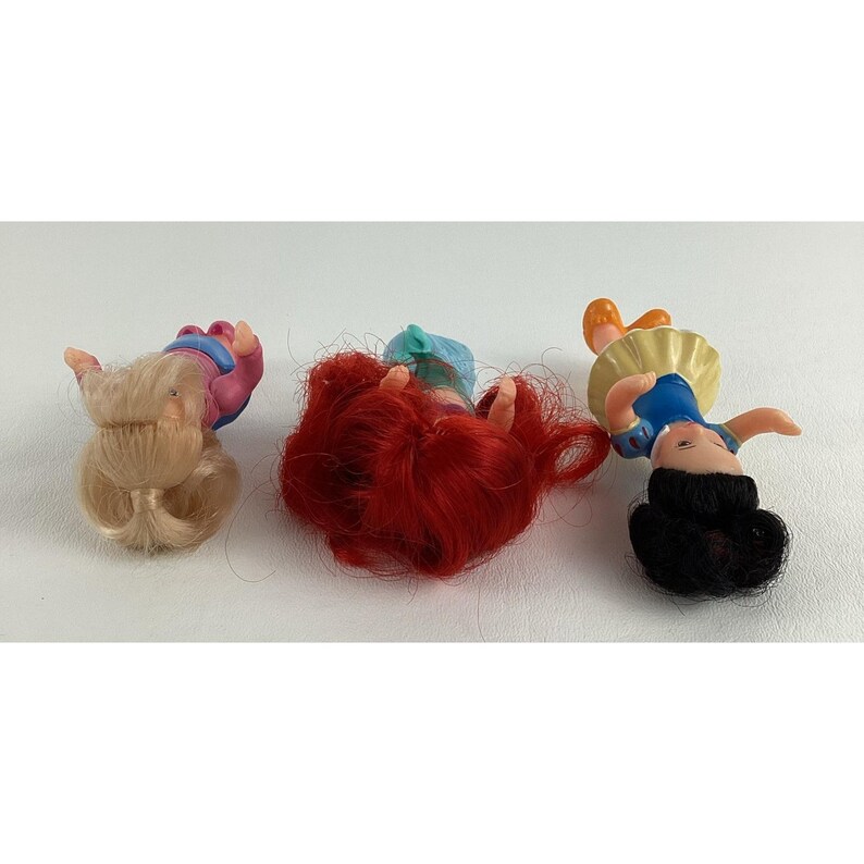 May include: Three small dolls with different hair colors and outfits are lying on a white surface. One doll has blonde hair and a pink and blue outfit. Another has red hair and a blue and green outfit. The third doll has black hair and a blue and yellow outfit.
