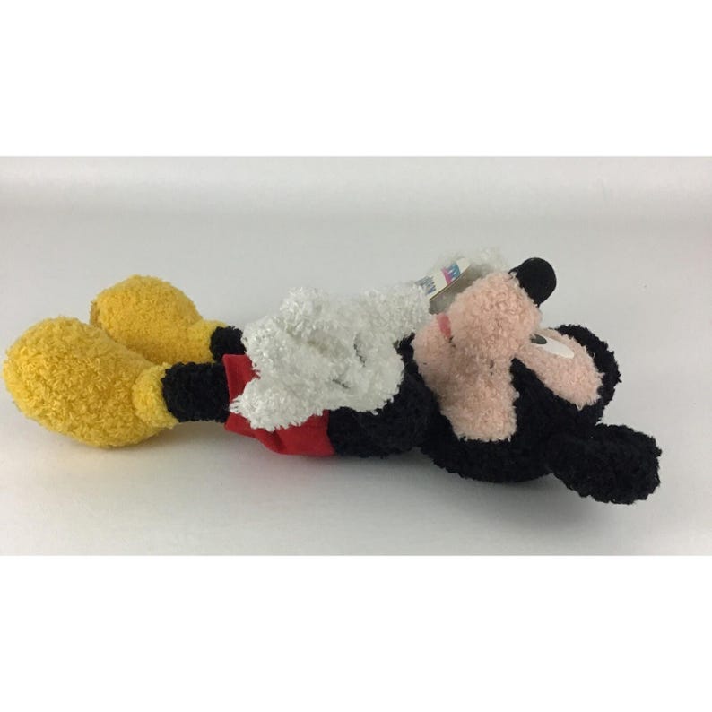 May include: A plush Mickey Mouse toy lying on its side. The toy features black fur, a pink face, white gloves, yellow feet, and red shorts. A tag is attached to the toy. The toy is on a white background.