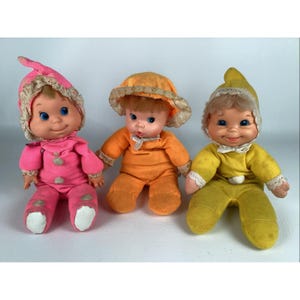 May include: Three vintage baby dolls in pink, orange, and yellow outfits. Each doll has a painted face with blue eyes and a smiling expression. The dolls wear matching hats with lace trim and are in a seated position.