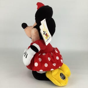 Pu&ograve; includere: Una bambola di peluche di Minnie Mouse, con un vestito rosso a pois bianchi, orecchie nere e un fiocco rosso abbinato. La bambola ha guanti bianchi e scarpe gialle. Un'etichetta Disney &egrave; attaccata.