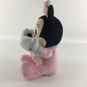 Op de afbeelding: Een roze pluche Minnie Mouse speelgoed dat een roze en groene outfit draagt met een witte strik. Het speelgoed zit met opgeheven armen en houdt een groen-wit boek vast.