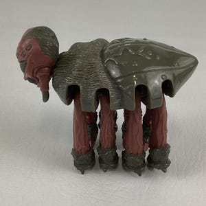 K&ouml;nnte beinhalten: Eine Kunststoff-Spielfigur in Form eines Wesens mit dunkelrotem Kopf und mehreren Augen, einem strukturierten grauen R&uuml;ckenpanzer sowie acht langen, dunkelroten Beinen mit grauen F&uuml;&szlig;en. Die Figur hat ein segmentiertes Aussehen mit gl&auml;nzendem Panzer.